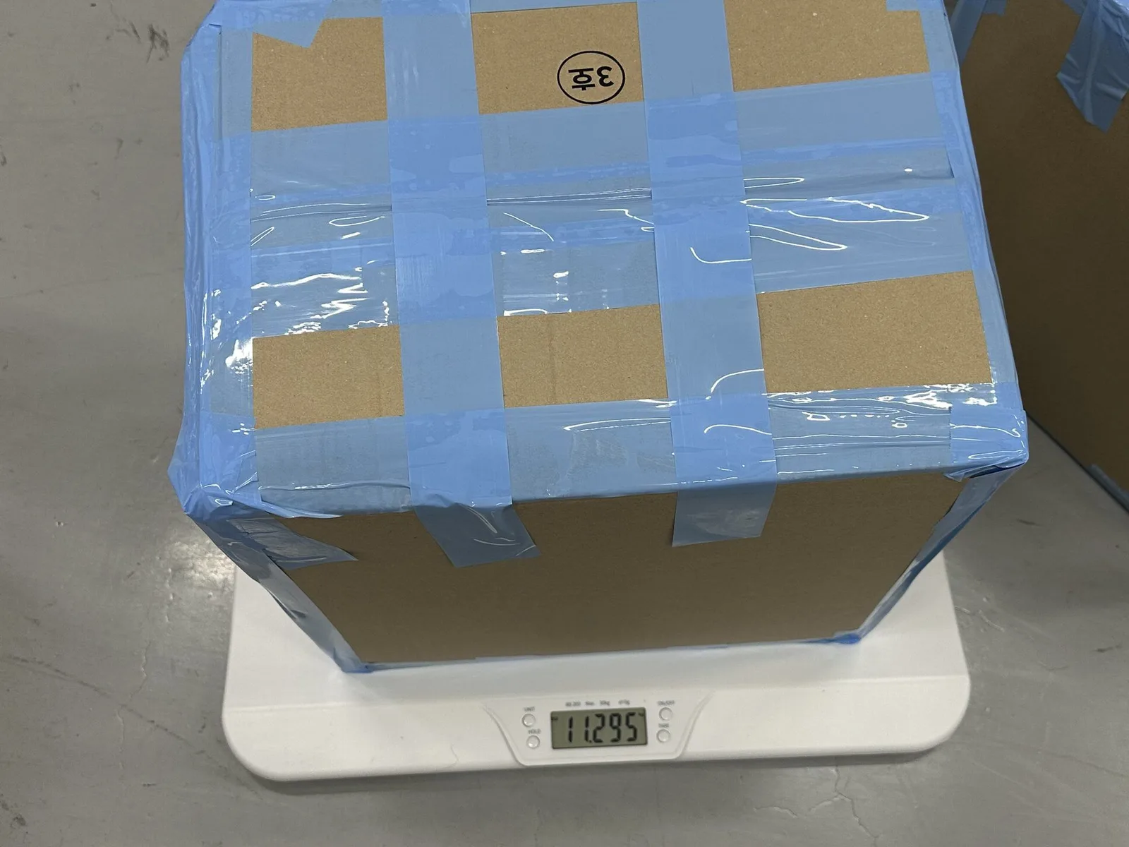 Sealed K-pop shipment carton being weighed on a digital scale